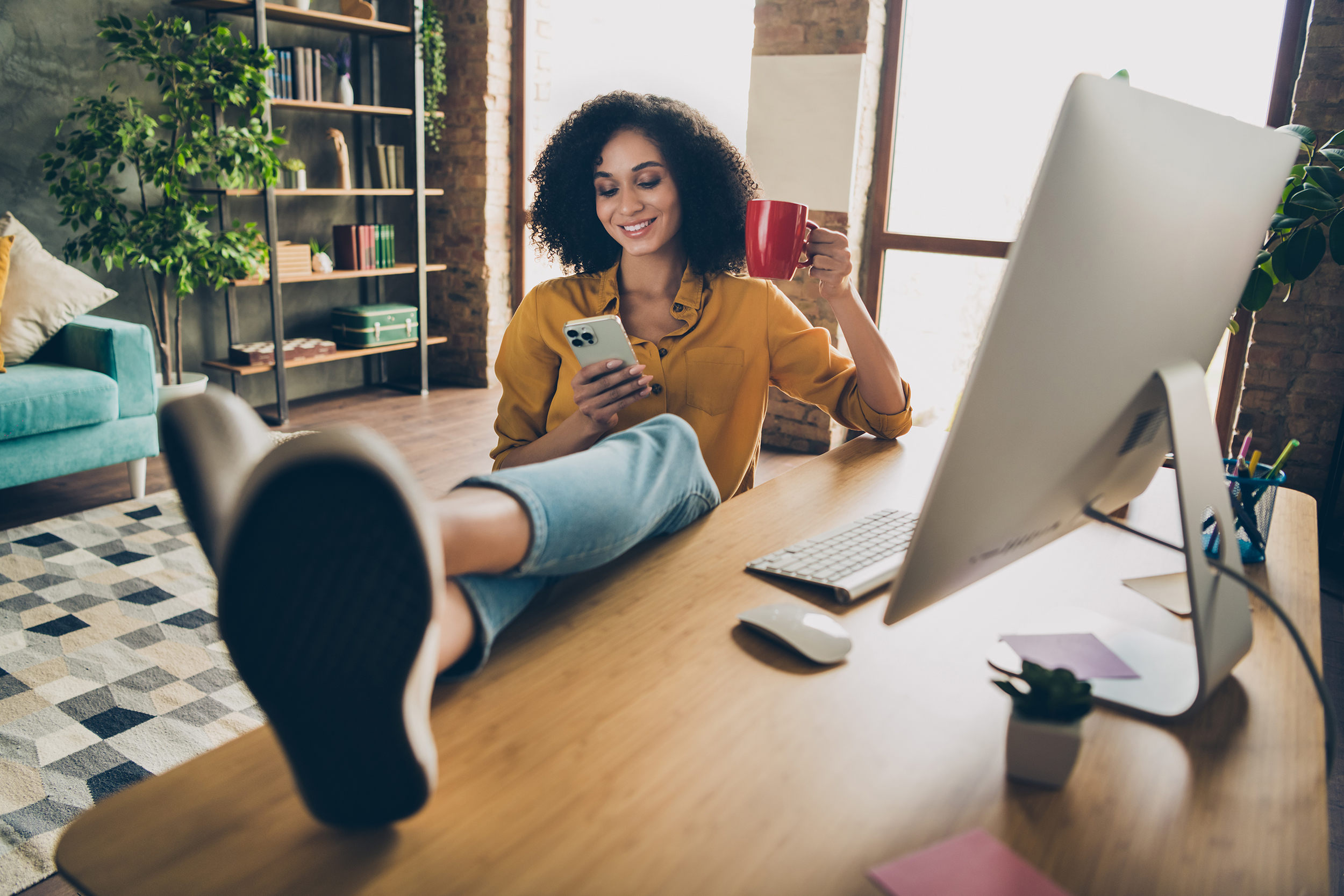 A woman, smiling, is casually reclining with her feet up in a modern, well-lit home office. She's holding a smartphone in one hand and a red mug in the other, while a desktop computer is in front of her. She appears relaxed and engaged, possibly taking a break to interact with her social media followers, enhancing her social media engagement, and embodying a sense of comfort and productivity.