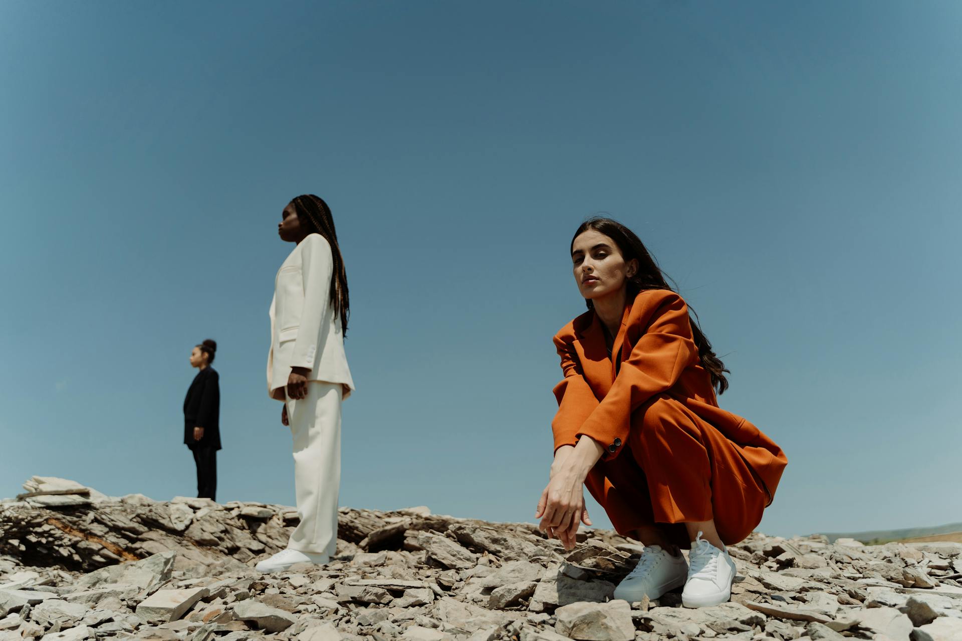 Three individuals in professional attire stand on a rocky terrain under a clear blue sky, embodying the concept of 'the zone.' The woman in the foreground, dressed in an orange suit, squats on the rocks, exuding focus and determination. The other two, a man in a black suit and a woman in a white suit, stand in the background, facing away, representing the discipline it takes to stay in 'the zone' amidst distractions