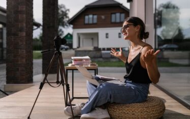 An enthusiastic content creator engaging with her audience through a live video session outdoors, with a professional camera set up on a tripod and a laptop on a small table, embodying the spirit of DIY personal brand content creation.