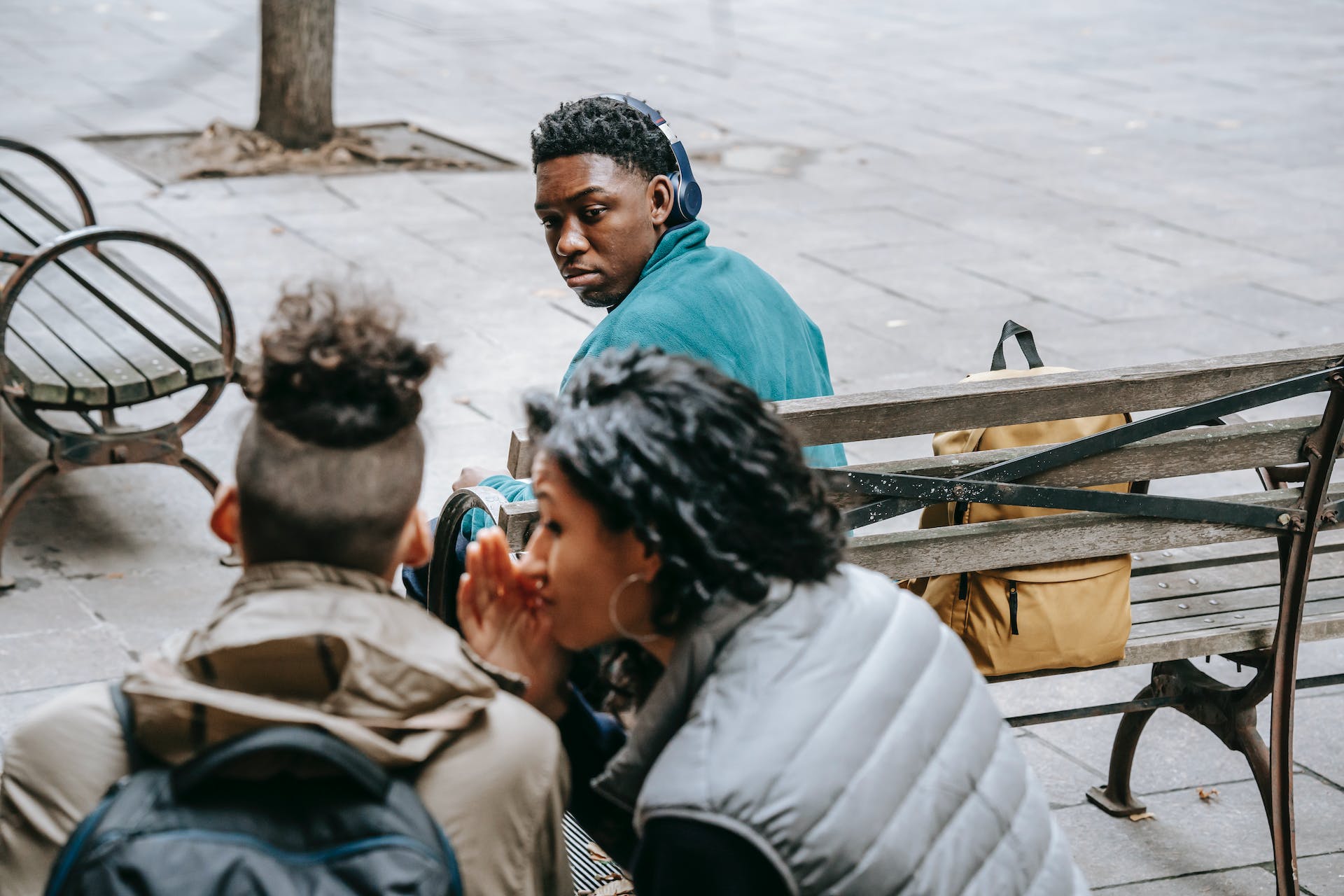 Three people seated on a public bench, with two engaged in a close conversation and the third person, wearing a teal hoodie and headphones, looking pensive as if reflecting on the conversation, encapsulating the theme of 'Responding to Criticism.