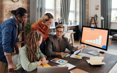 Four professionals engaged in brand building growth strategy, analyzing graphs on a computer monitor in a well-lit, modern workspace.