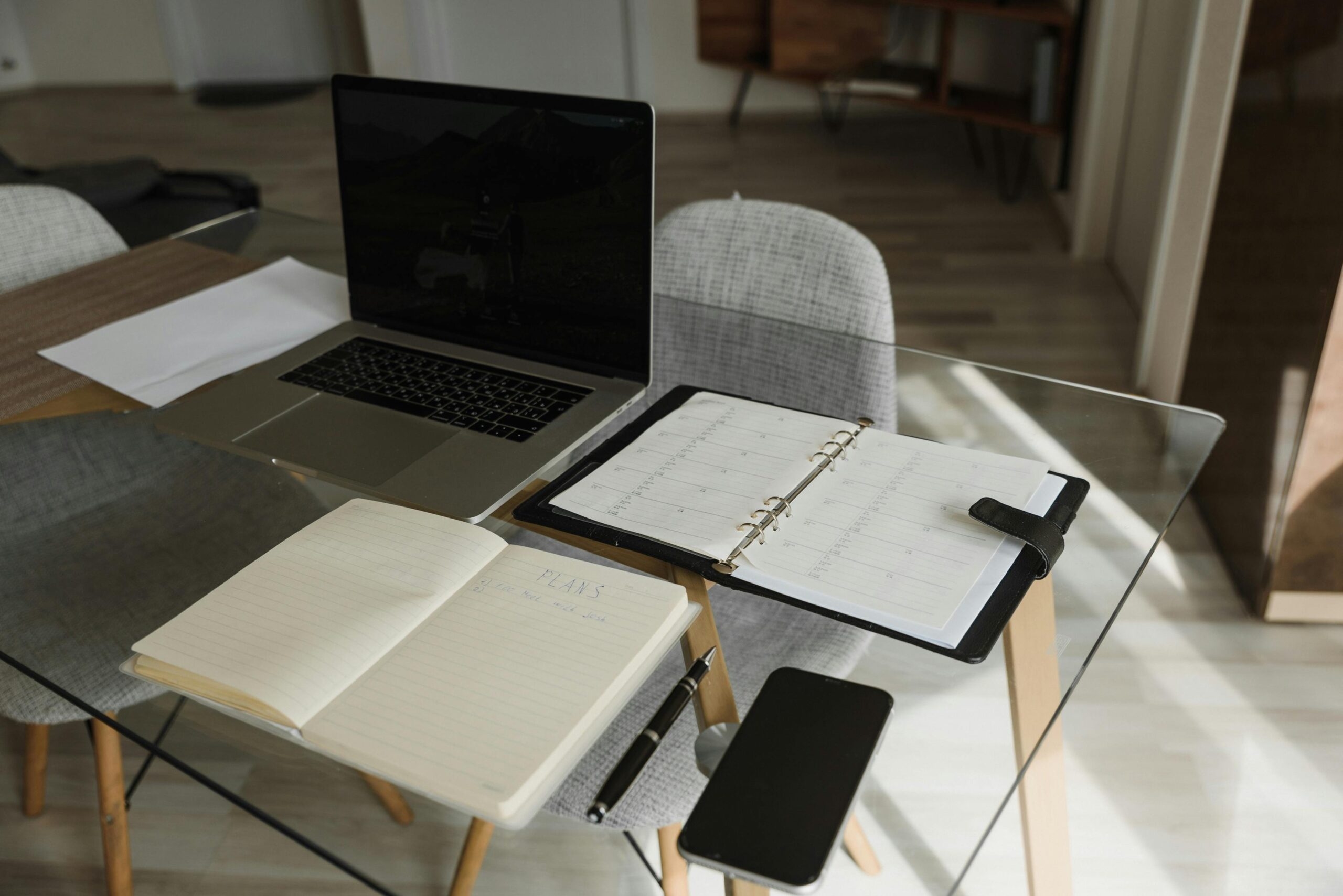 A modern workspace featuring an open laptop on a glass table, with a planner opened to a blank page alongside a pen, all ready for planning. A smartphone rests nearby, symbolizing the integration of digital and traditional planning. This serene setting invites contemplation on the question: What will you do to make this a successful year?