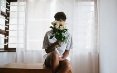 A person sitting serenely on a wooden stool, holding a bouquet of fresh, blooming flowers close to their face, symbolizing a moment of self-care and the practice of intentional love.