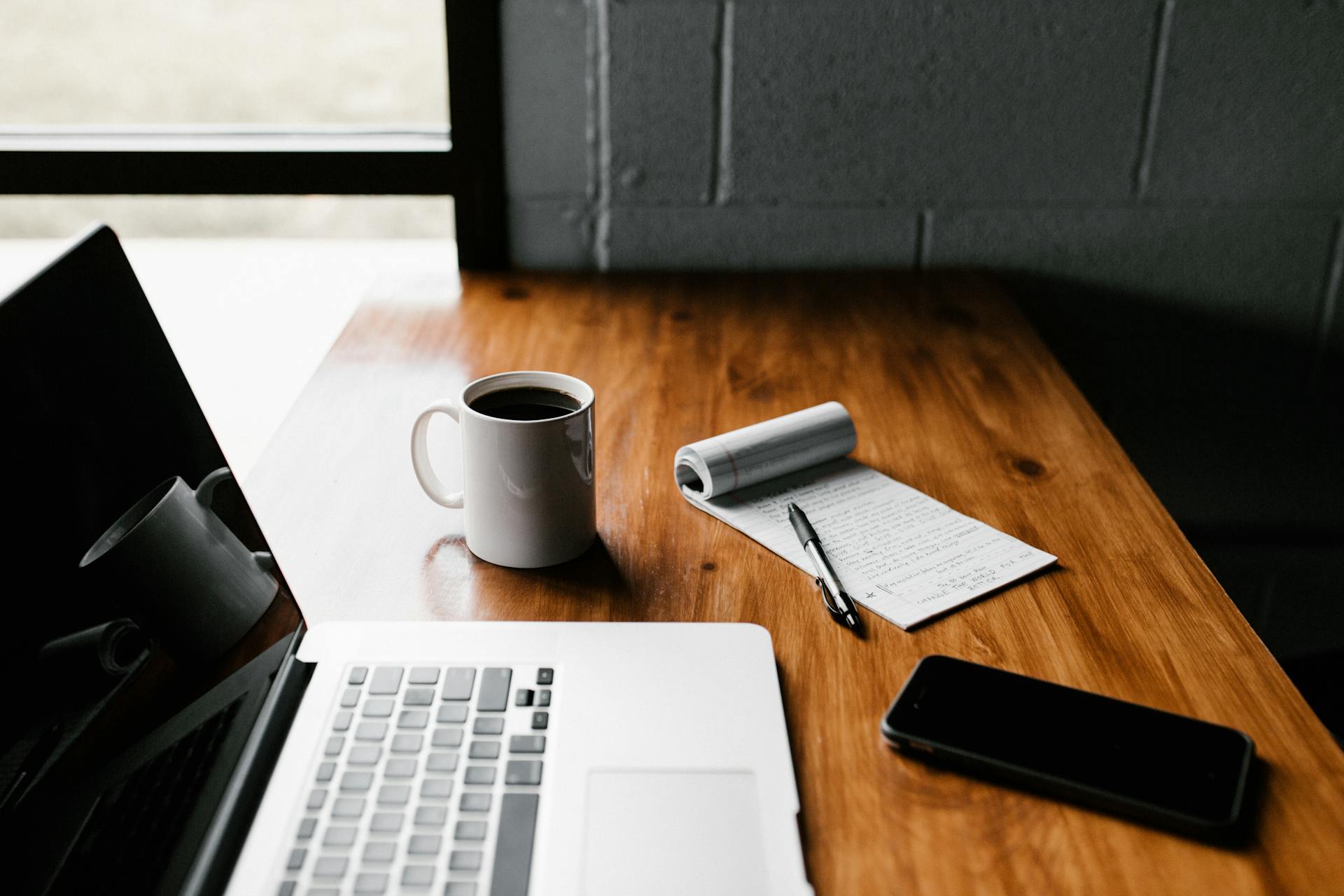 Wooden desk featuring a laptop, a cup of coffee, a notepad with a pen, and a smartphone, symbolising Faith and Work Balance.