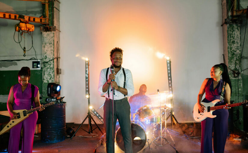 Black audiences support local artists performing live with guitars, drums, and stage lighting in an urban creative venue.