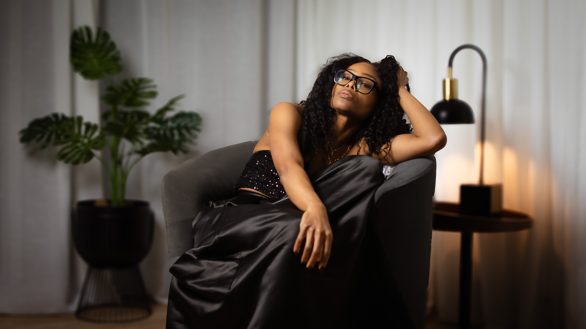 Bess Obarotimi seated in a softly lit room, wearing a black outfit and glasses, embodying calm confidence and deep reflection on spiritual transformation work.