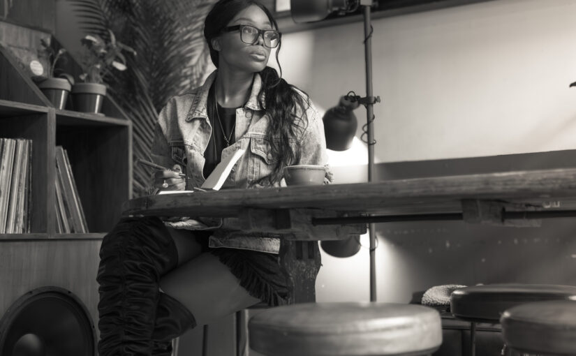 Black-and-white portrait of a woman seated at a table, writing in a notebook, looking away thoughtfully in a café setting.