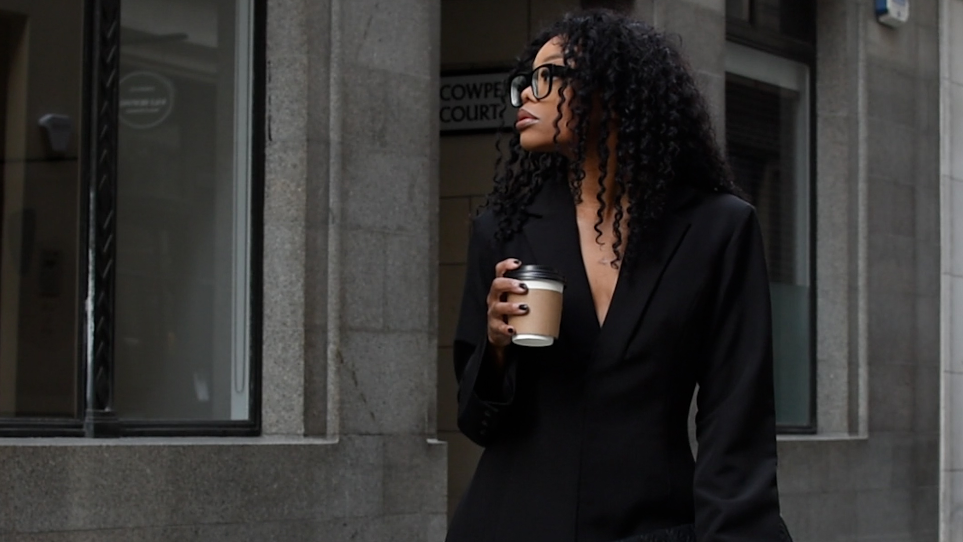 Woman in a black suit holding coffee outside an office building.